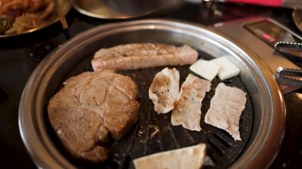 Hand uses tongs to turn garlic on grill with pork, steak, and vegetables, warm lighting