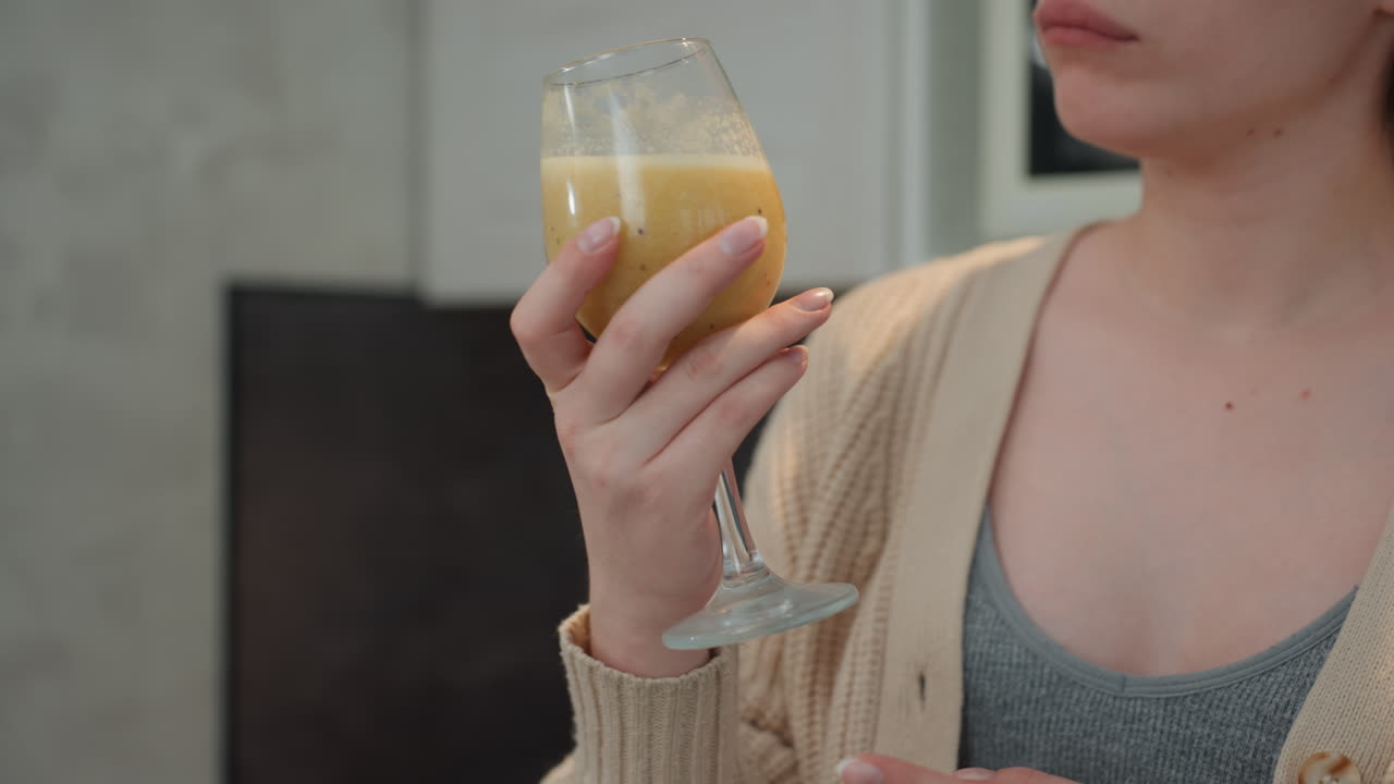 Mujer caucásica embarazada inspeccionando vaso de batido, estudiante de nutrición examinando con vacilación la textura y el aroma, mano cuidadosa en el tallo, mirada atenta evaluando seguridad y sabor, fondo de cocina tenue.