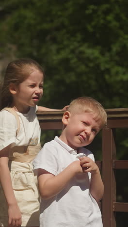 bored little children stand on bridge near waterfall in wild park. boy and girl tired of long walk to cascade on mountain river on summer vacation