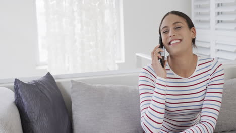 Happy-biracial-woman-sitting-on-sofa-talking-on-smartphone,-copy-space,-slow-motion