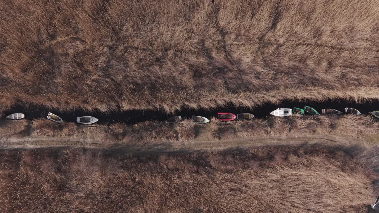 Flygbild uppifrån av små färgglada båtar förtöjda i en smal kanal omgiven av tät torr vass. En slingrande grusväg på landsbygden löper parallellt med vattenvägen.