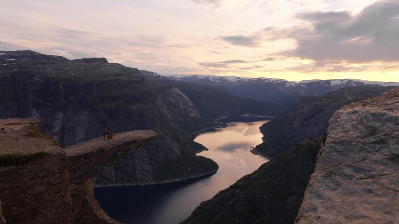 Person with dog on cliff at sunset, overlooking Norwegian fjord and vast mountains, Trolltunga