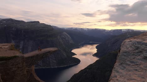 person with dog on cliff at sunset, overlooking norwegian fjord and vast mountains, trolltunga