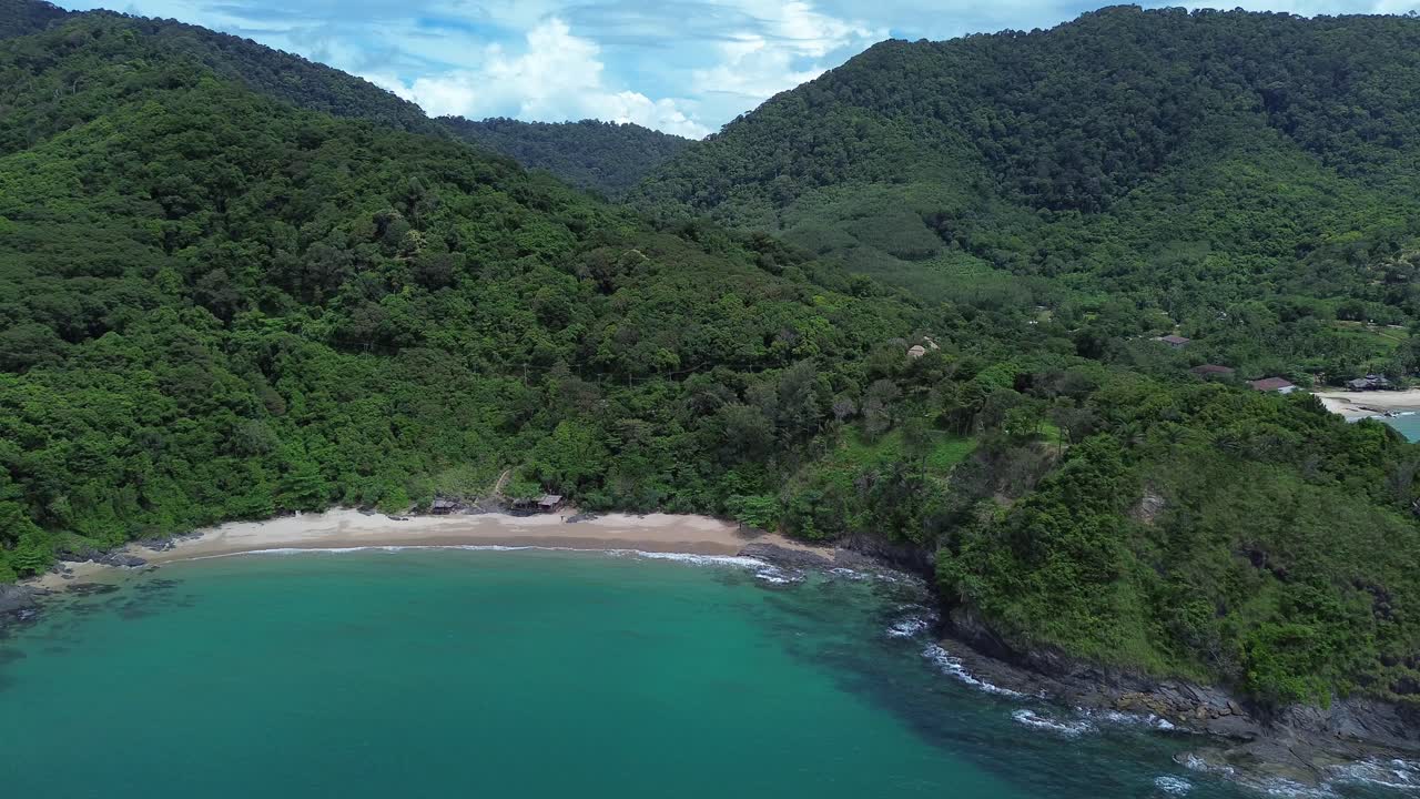 Aerial drone view of a remote Koh Lanta beach with lush green hills, dense forest and a few scattered coastal bungalows, showing turquoise water and peaceful tropical island scenery without crowds