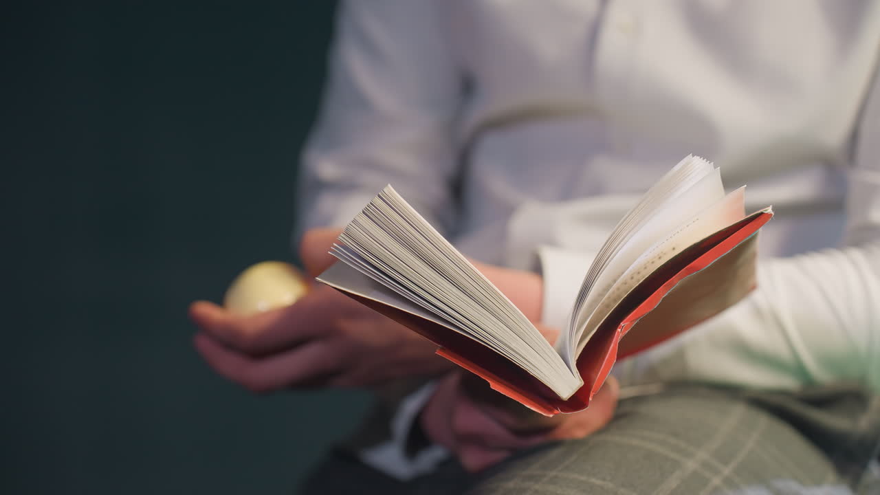 toma de primer plano de la mano de una persona sin rostro sosteniendo un libro abierto mientras hace rodar una pelota de billar en una mano. borrón suave en el fondo, creando un estado de ánimo artístico y reflexivo con enfoque en los gestos de la mano