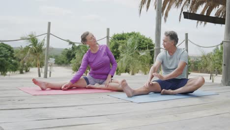 happy senior caucasian couple practicing yoga sitting on beach sun deck, in slow motion