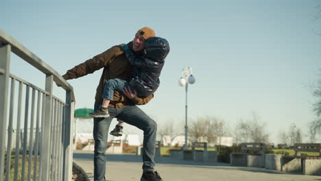 close up of father carrying his son as he hold the rail with his right hand and hang one leg in the air, they both are laughing and happy as he plays and dangles