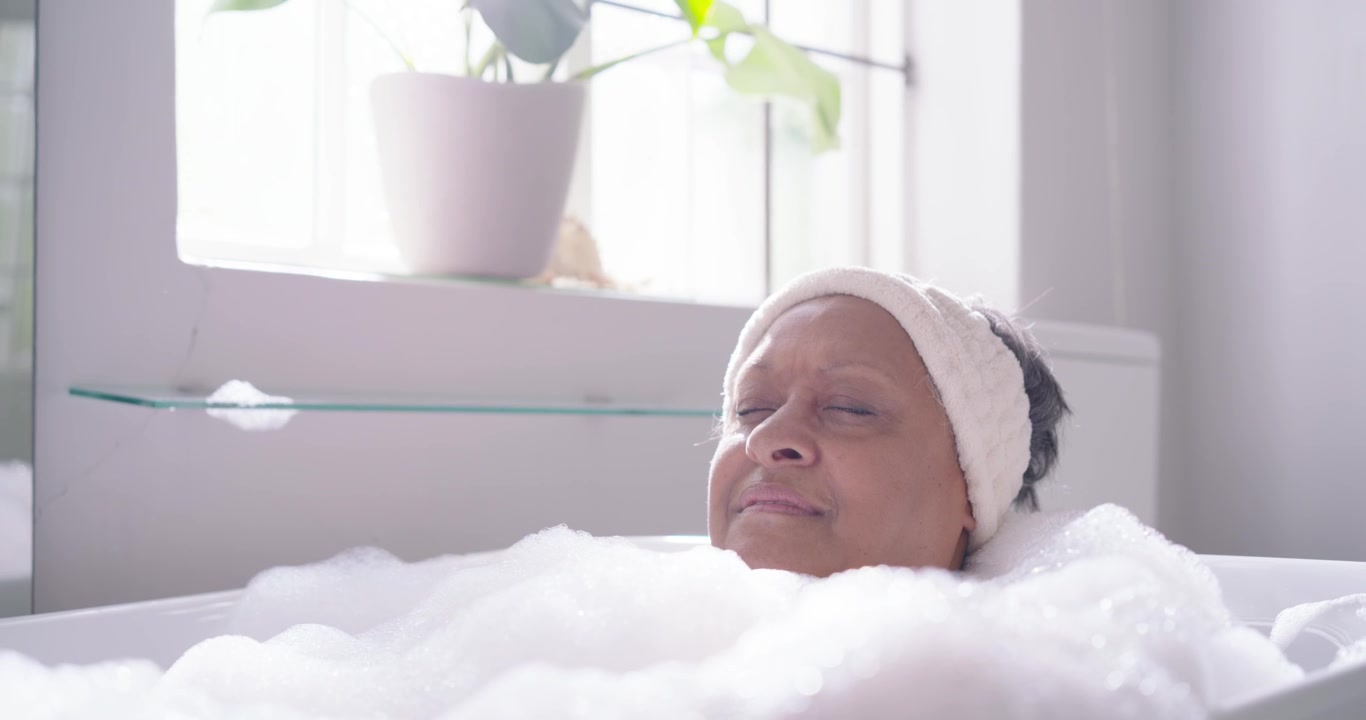Senior woman reclining in bathtub with foam, relaxing while camera widening revealing plant