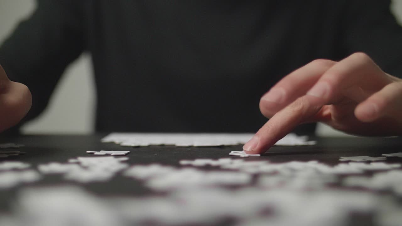 Slow Motion: Young man's hands taking a puzzle piece, trying to solve a puzzle, while more pieces are laying around on a dark table. Shot in 4K, Slow Motion with Shallow Depth of Field