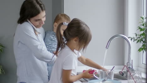 mother holds her little baby and talks on the phone, while her little daughter washes the dishes