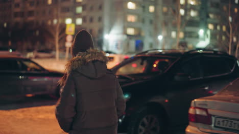 first-person view of a woman in a winter coat with a fur hood, walking towards a black car at night in a parking lot. the background features parked cars and illuminated buildings