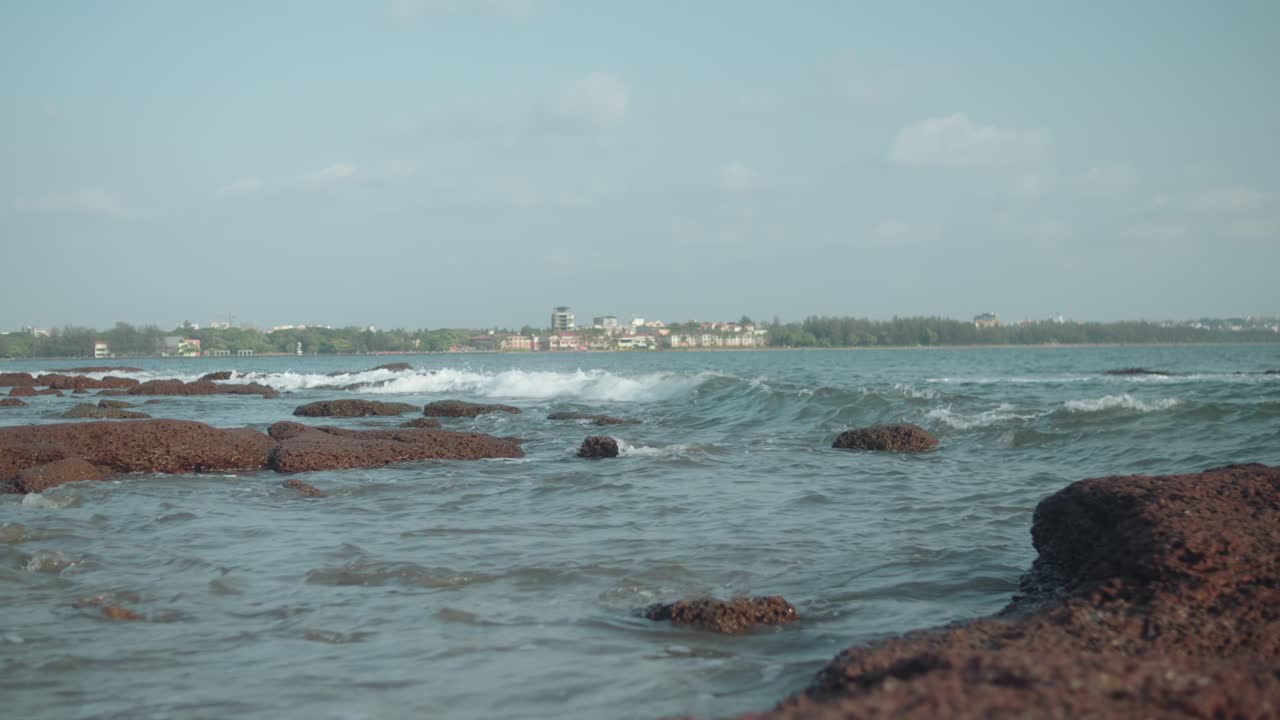 Scenic shot from Chicalim Bay captures waves rolling over reddish laterite rocks in the foreground, with distant buildings of Vasco da Gama across blue water under a clear Goan sky