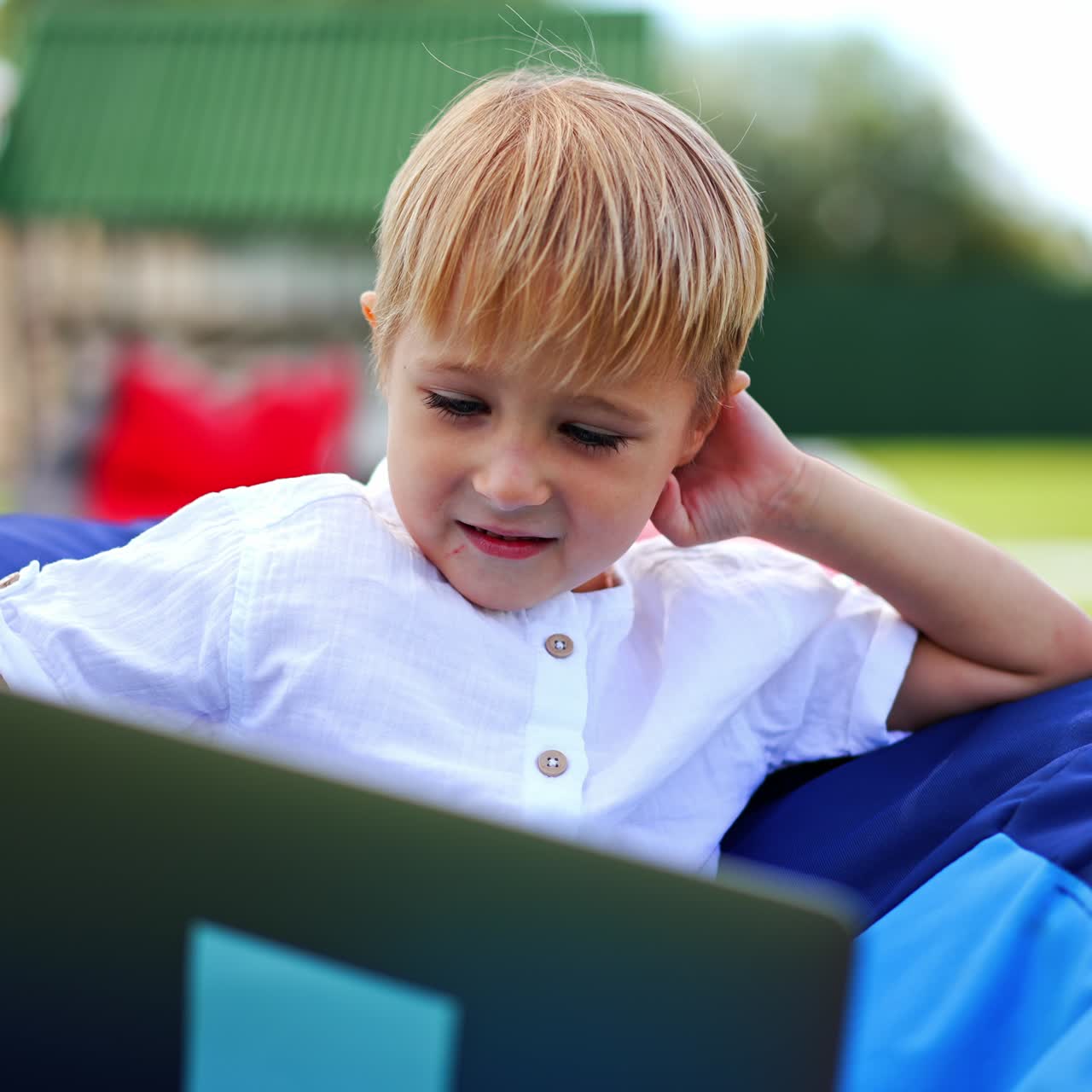 Lovely blond kid using gadgets outdoors. Nice little boy sitting in bean bag chair watching videos on laptop smiling. Close up. Blurred backdrop