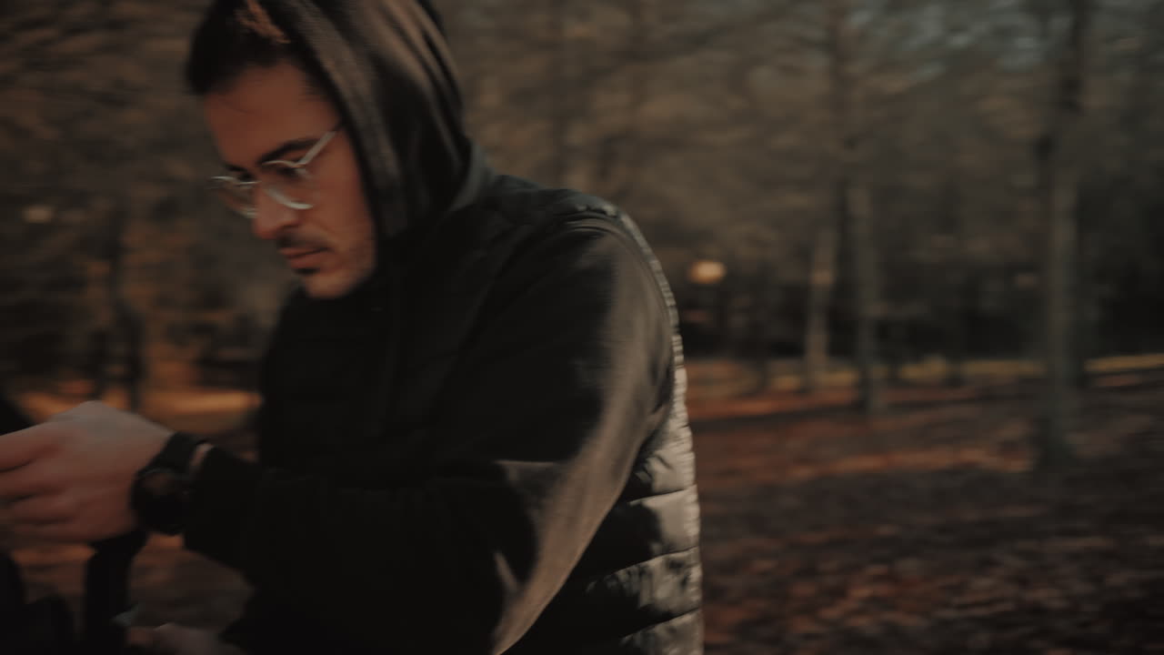 joven videoasta caucásico enfocado con anteojos, chaleco negro y capucha, trabajando al aire libre y grabando video al atardecer usando una cámara dslr sin espejo montada en un cardán estabilizador motorizado