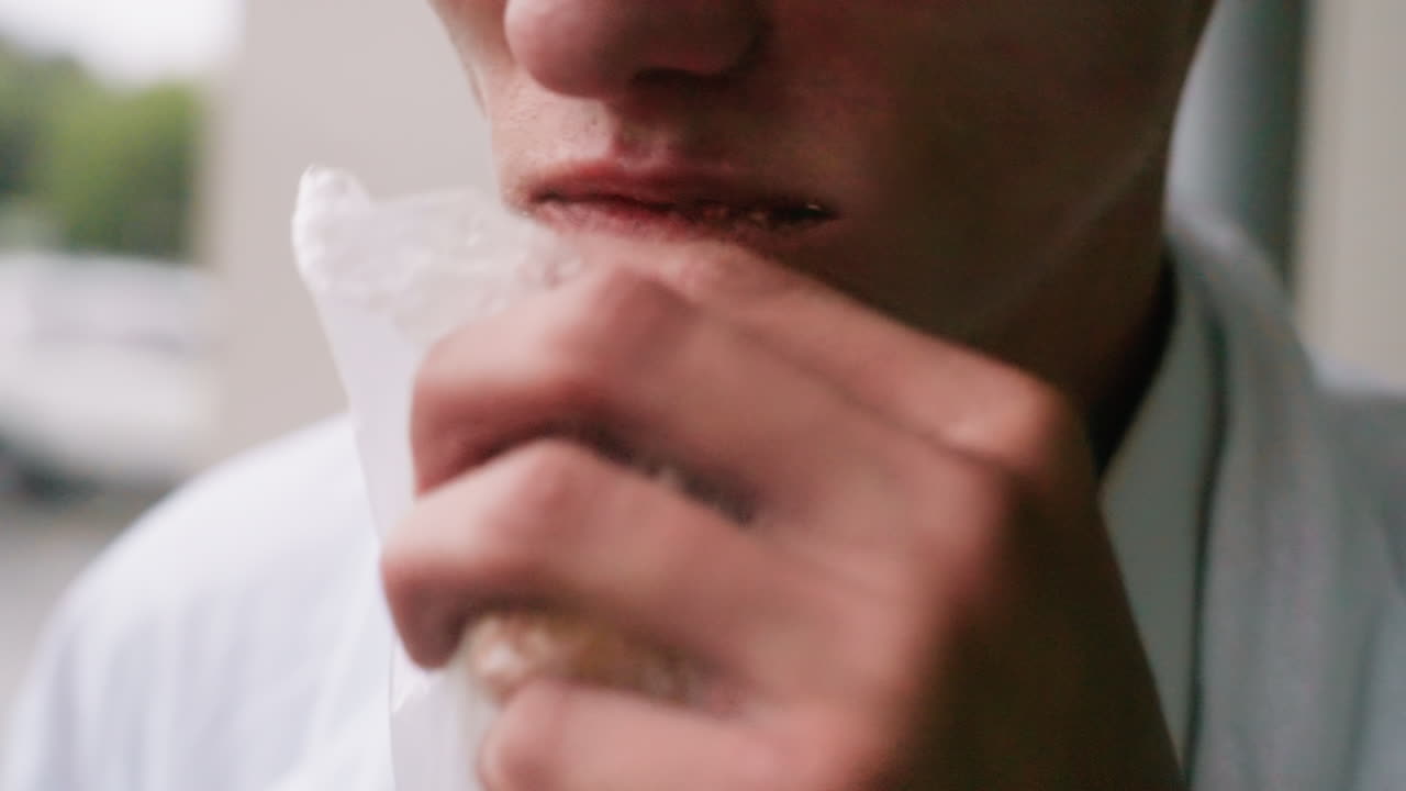 Close up view of young man in white coat holding bitten pastry wrapped in plastic bag while chewing snack during outdoor break, showing simple daily moment of eating with casual detail