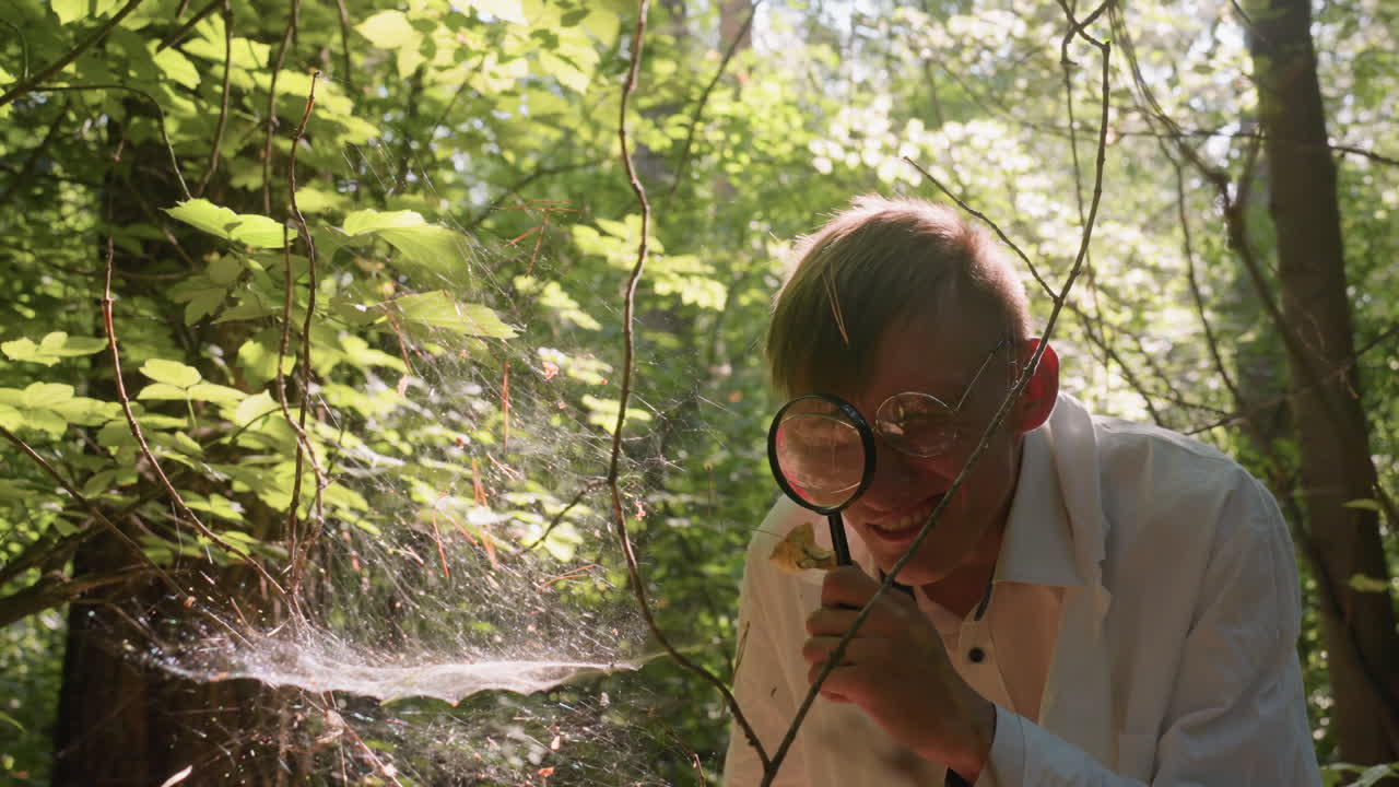 Young man in white coat carefully observing spider web with magnifying glass in dense forest, focusing on delicate cobweb structure illuminated by sunlight for ecological exploration and scientific observation