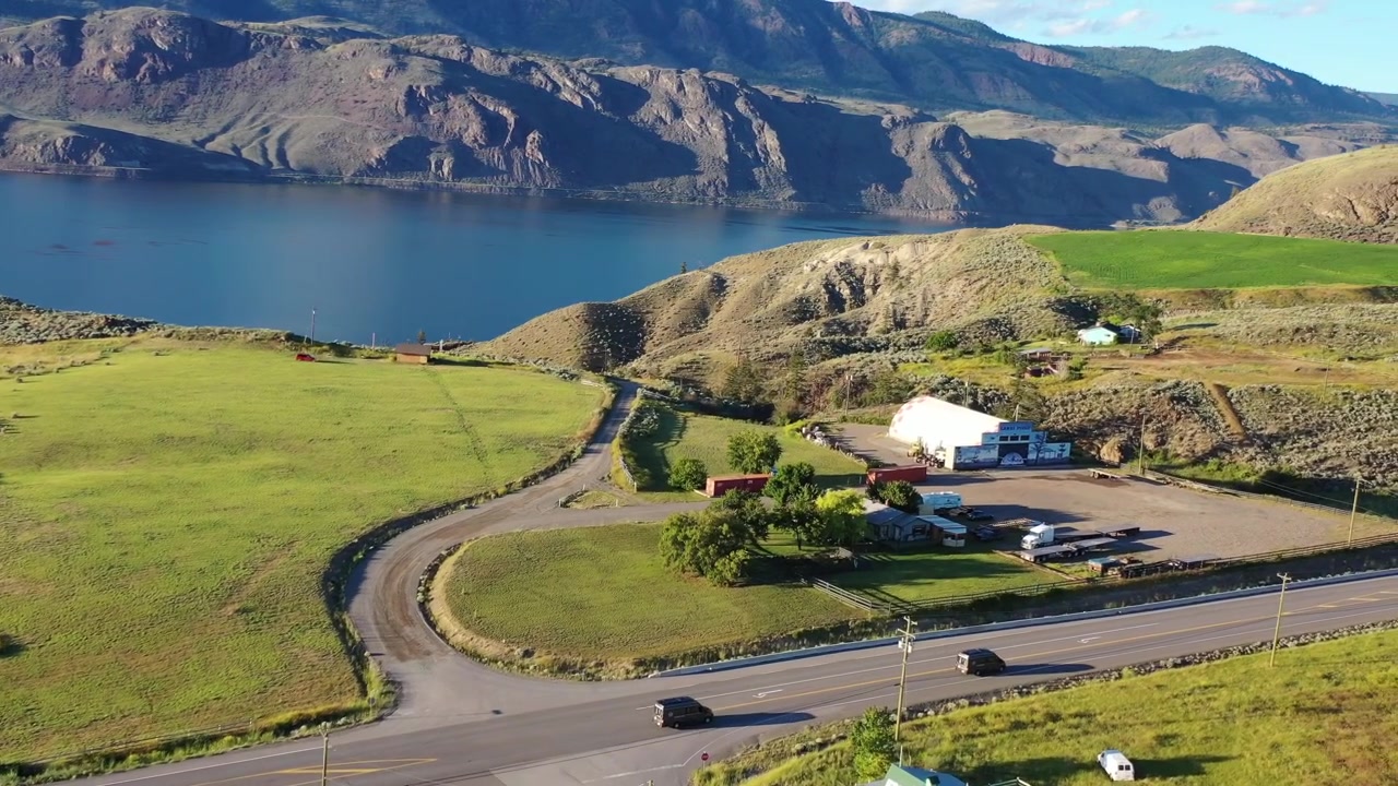 Ripresa con drone dall'alto di due furgoni che viaggiano insieme su una strada asfaltata attraverso dolci colline e campi verdi. Una scena cinematografica di un viaggio on the road in stile "van life" nell'entroterra della Columbia Britannica sotto un sole pomeridiano splendente.