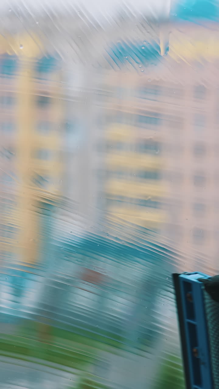 Washing a window in the city. Wet window is cleaning with special brush on blur background of multi-storey buildings. Vertical video