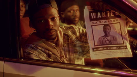 young black man posing with wanted poster in car illuminated by police lights