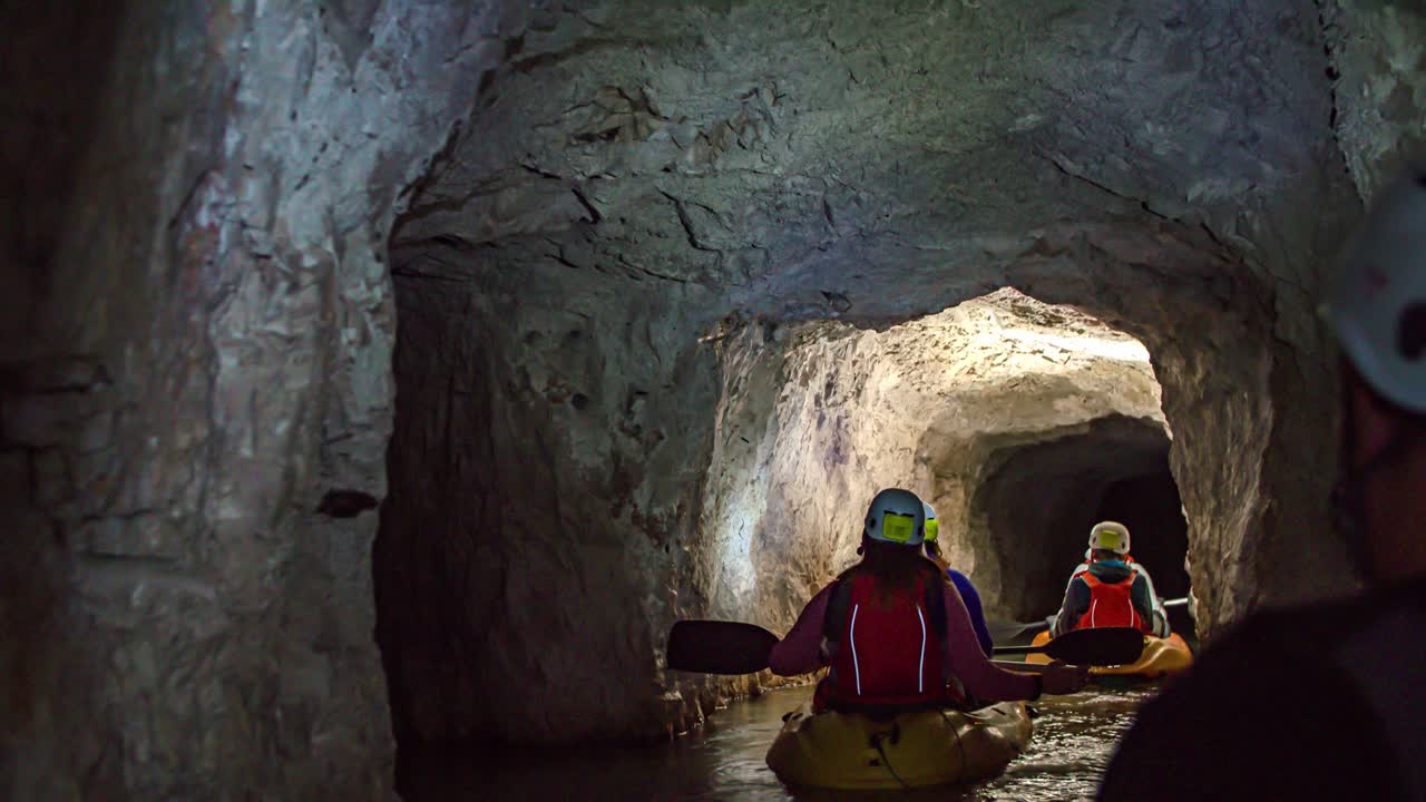 Tourists travel on two kayaks inside underground narrow mining tunnel in dark rocky cave, behind view