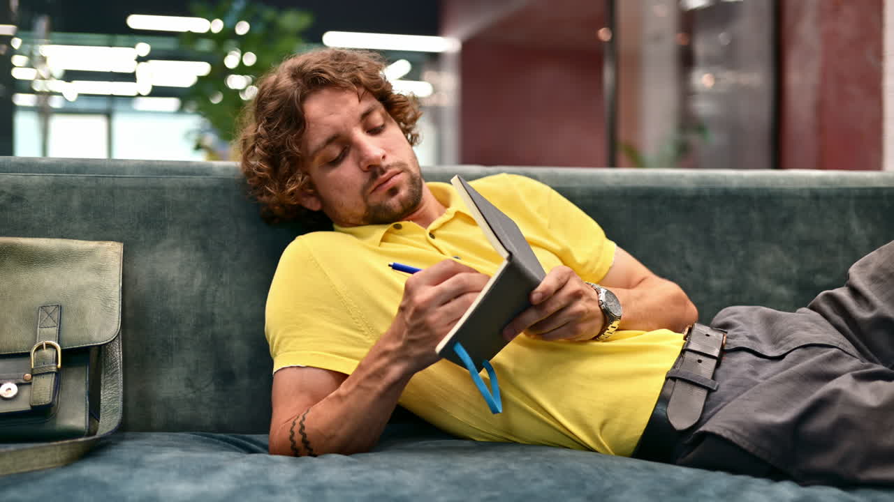Man in yellow shirt lying on a couch and writing in a notebook