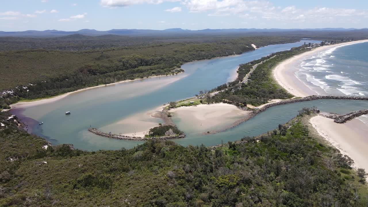 Aerial view of a peaceful coastal inlet with a jetty, where a river flows into the ocean, surrounded by lush greenery and sandy shores, showcasing nature's beauty