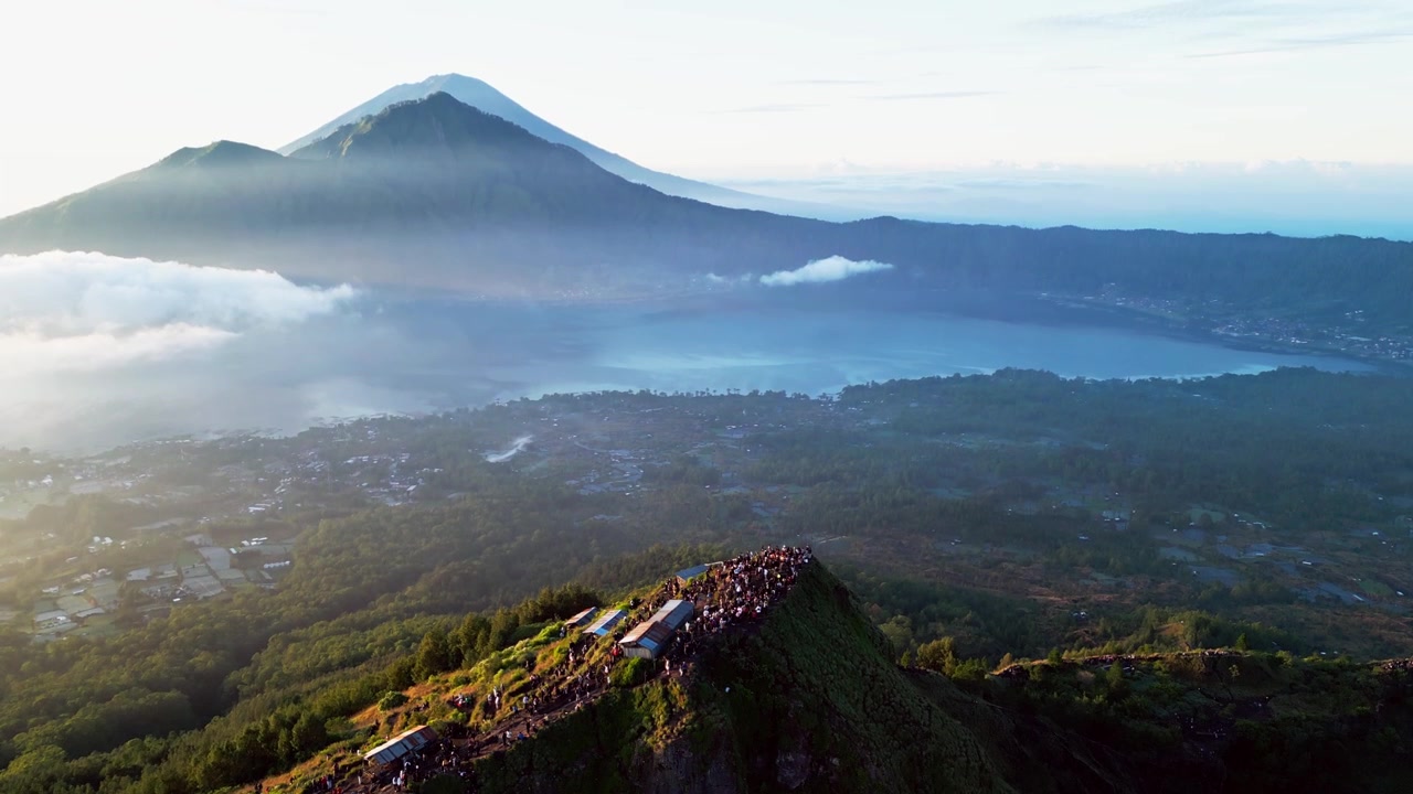 Nascer do sol sobre uma estreita crista de montanha com caminhantes em trilha exposta no cume, cena contraluz com névoa revelando lago de cratera e pico vulcânico em Bali, avanço com tomada de drone revelando em inclinação para cima