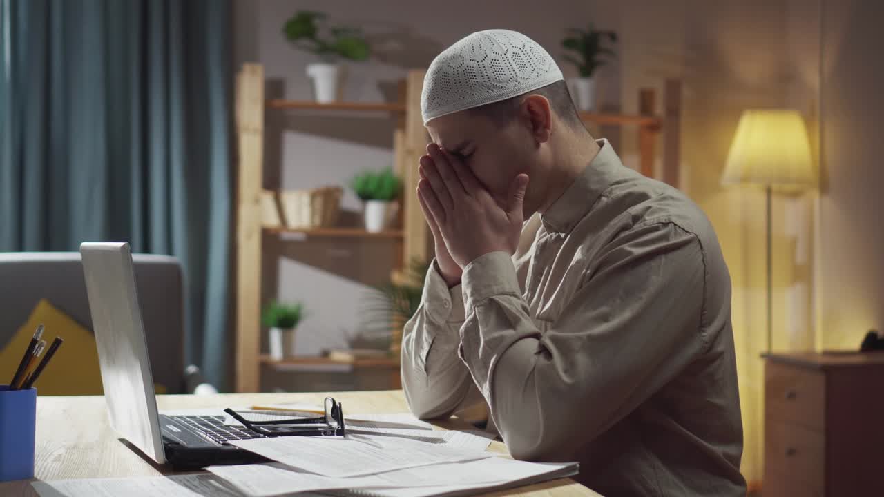An agitated muslim man in glasses and a skull-cap sits in the living room at a laptop and counts finances