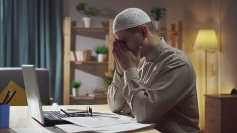 an agitated muslim man in glasses and a skull-cap sits in the living room at a laptop and counts finances