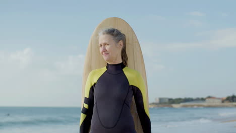 front view of a cheerful senior woman standing on seashore with surfboard and looking at the camera