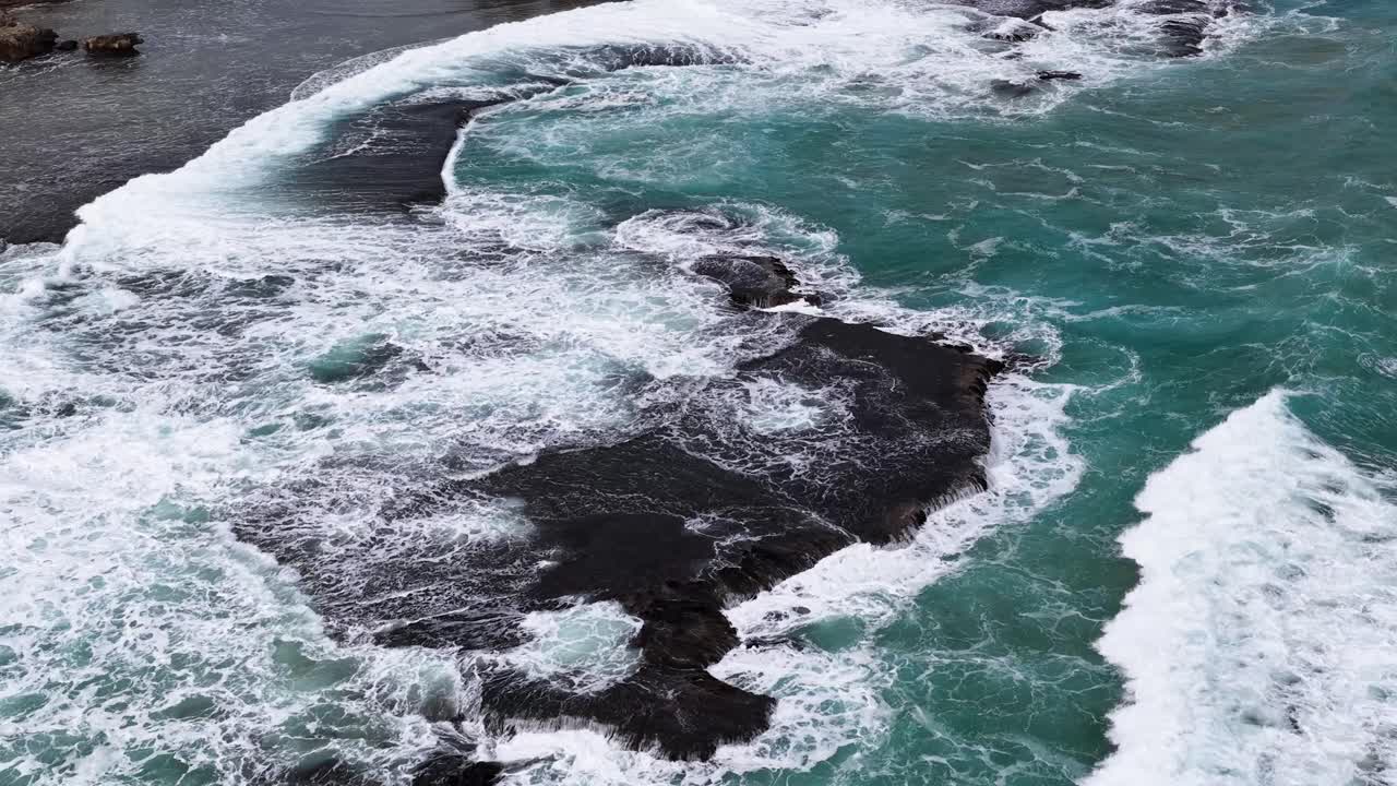 Overhead drone shot of turquoise waves breaking on dark coastal rocks, natural daylight, dynamic movement