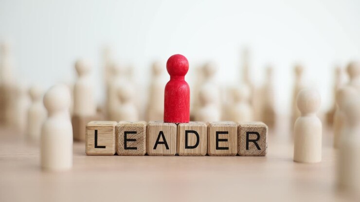 A red leader figure stands on "LEADER" blocks among other wooden figures, symbolizing leadership and prominence