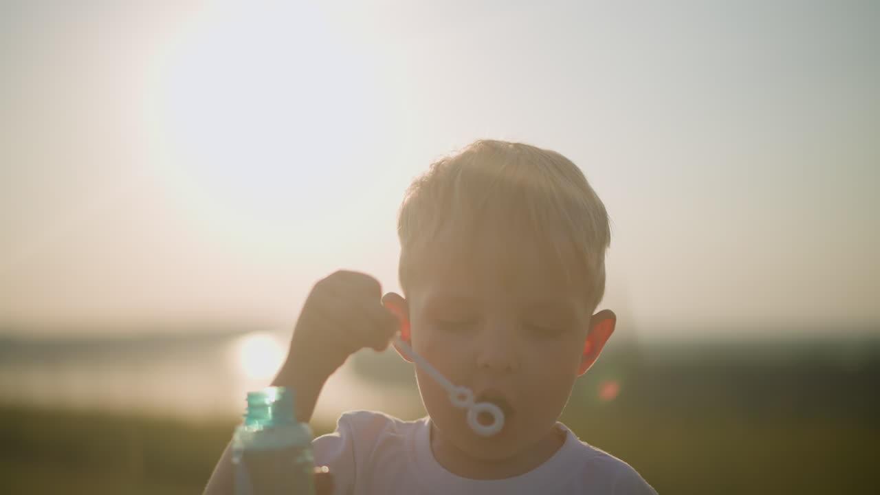 A little boy wearing a white shirt blows bubbles into the air at sunset, with a woman s hand holding the foaming bottle. The warm sunlight enhances the serene, joyful moment as the child focuses