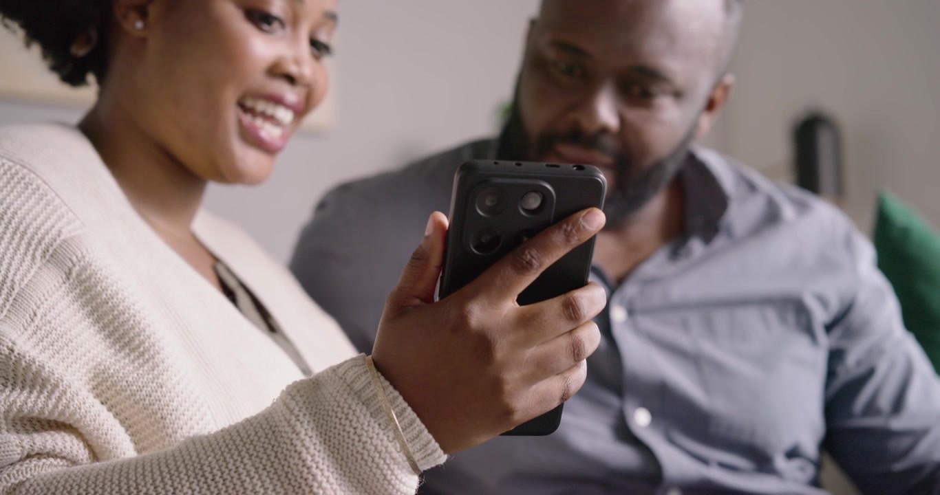 African couple sitting at home on sofa, woman tapping smartphone sharing content while man leaning