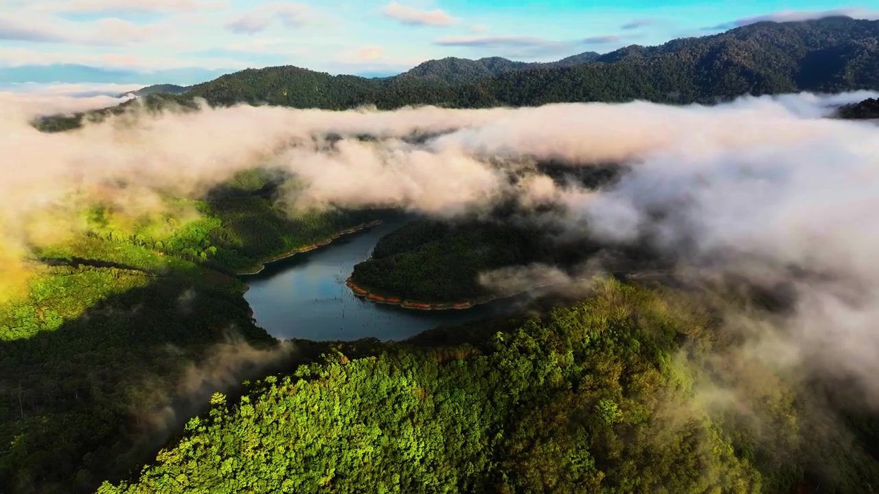 Atmospheric low clouds drift across lush green hills surrounding a calm body of water.