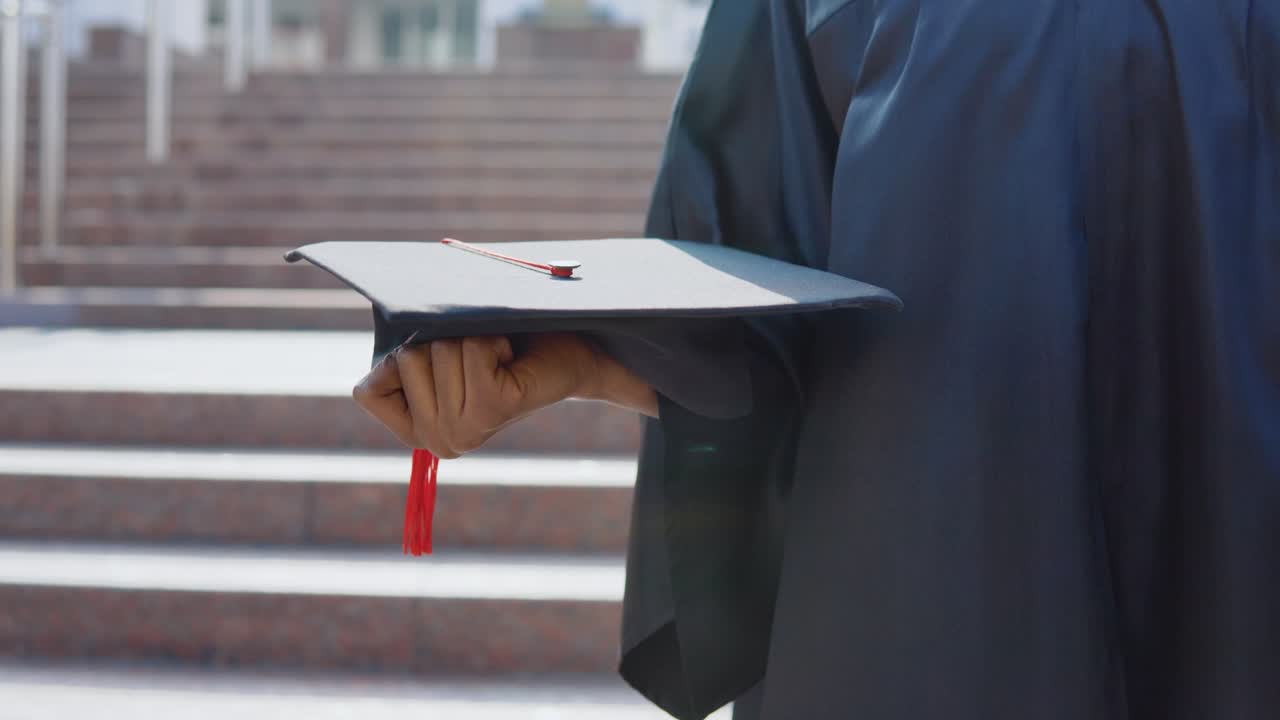 sombrero de maestro cuadrado horizontalmente en las manos de una mujer afroamericana graduada de la universidad en el fondo de las escaleras desde el exterior.