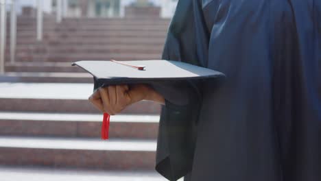 square master's hat horizontally in the hands of an african-american female university graduate on the background of the stairs from the outside.