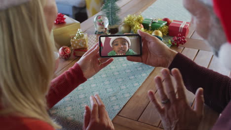 pareja caucásica con sombreros de santa usando un teléfono inteligente para una videollamada de navidad con la familia en la pantalla