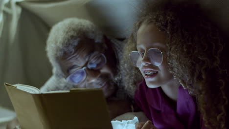 cute girl and her grandpa reading book, hiding under blanket