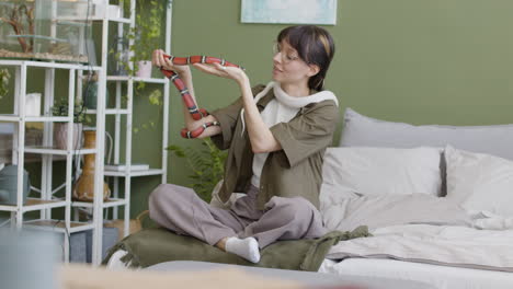 happy young woman holding her two pet snakes while sitting on bed at home 1