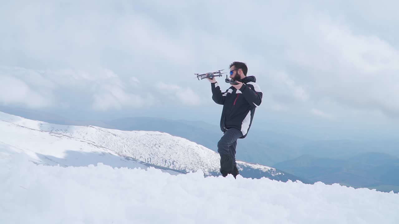 Uomo barbuto in piedi nella neve in cima a una montagna, che tiene un drone, fa una faccia buffa e scuote la testa