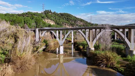 concrete bridge with arches reflected in calm river water, surrounded by trees and hills under a clear blue sky.
