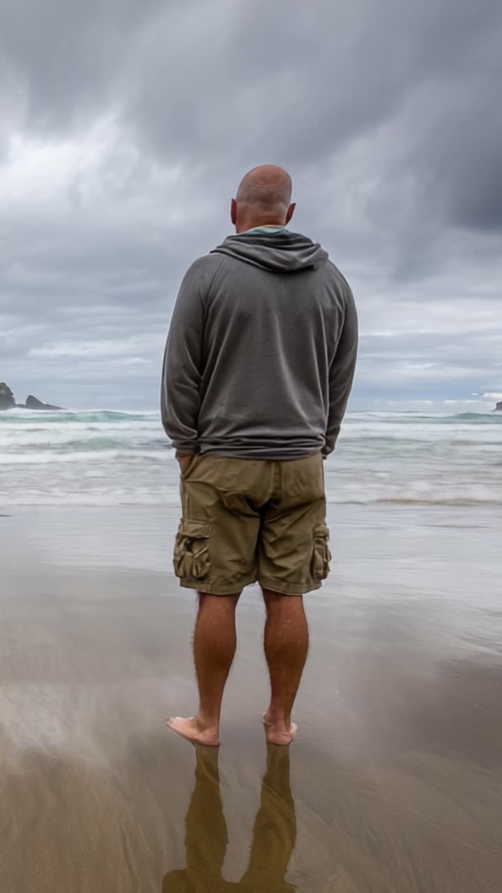 Un momento solenne sulla riva: un uomo sta in piedi da solo sulla spiaggia, contemplando le onde turbolente e i cieli minacciosi sotto un'atmosfera coperta.