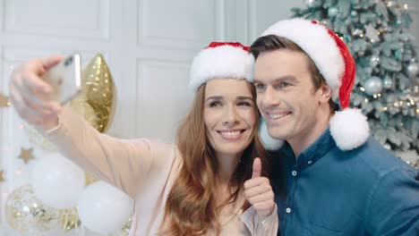 retrato de una pareja sonriente haciendo selfie con gorros de papá noel cerca del árbol de navidad.