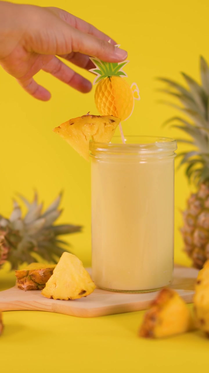 Una mano posiziona una fetta d'ananas su un frullato cremoso, circondato da frutta fresca, sotto una luminosa illuminazione da studio