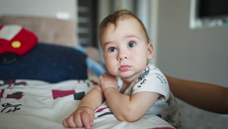 little sweet calm baby boy near the bed. adorable caucasian child looking into camera with interest. close up. blurred backdrop.
