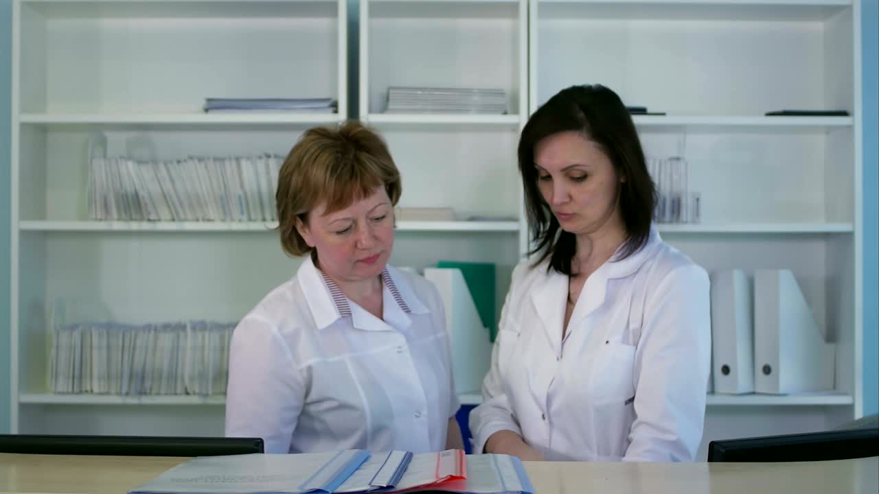 Dwie pielęgniarki porządkujące segregatory i dokumenty przy recepcji szpitalnej.