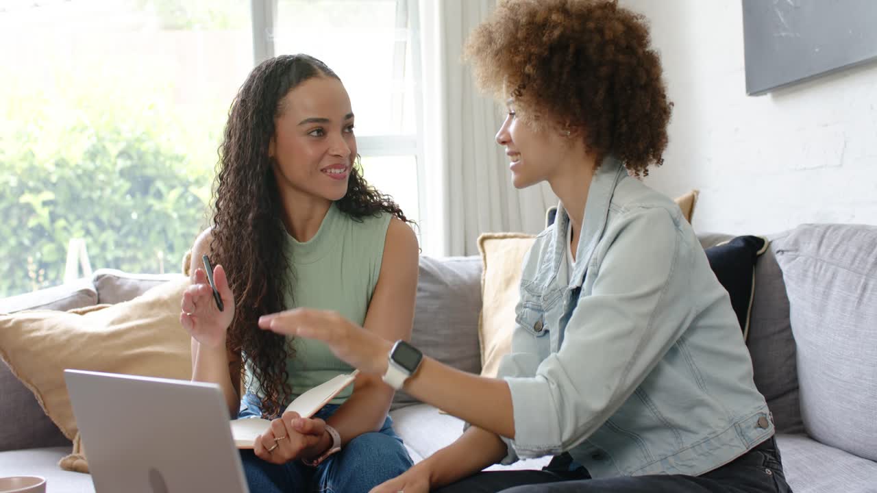 Mulheres afro-americanas trabalhando no sofá, uma apontando para o notebook, outra digitando no laptop, e depois fazendo um high-five.