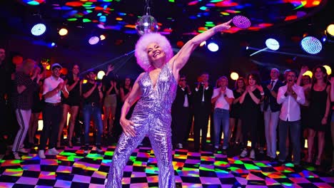 energetic senior woman with sparkling jumpsuit and white afro hair dancing on checkered dancefloor while audience is clapping hands in colorful nightclub