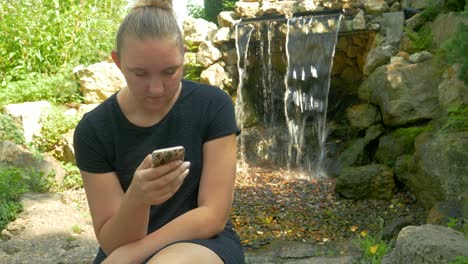 hermosa mujer en la naturaleza usa su dispositivo móvil mientras se sienta en un banco del parque junto a una cascada